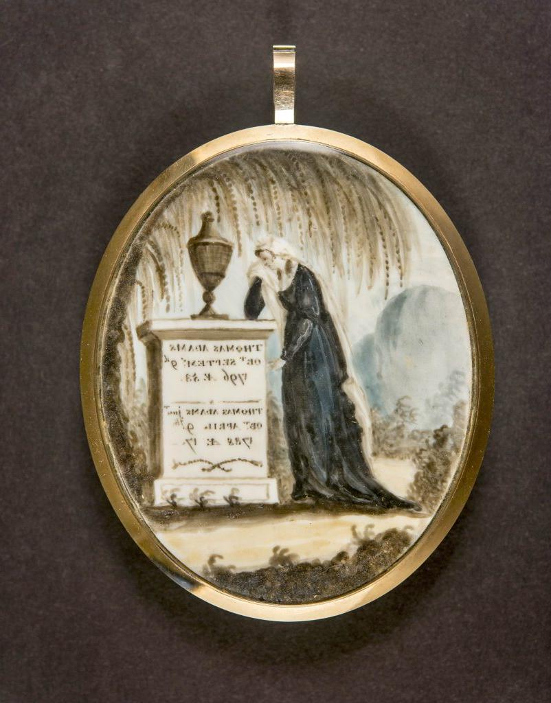 A color photograph of a painted image in an oval with gold metal around it making it into jewelry. The painted image is of a woman crying over a gravestone with an urn on top and a weeping willow tree above her. The gravestone has writing on it for Thomas Adams.  