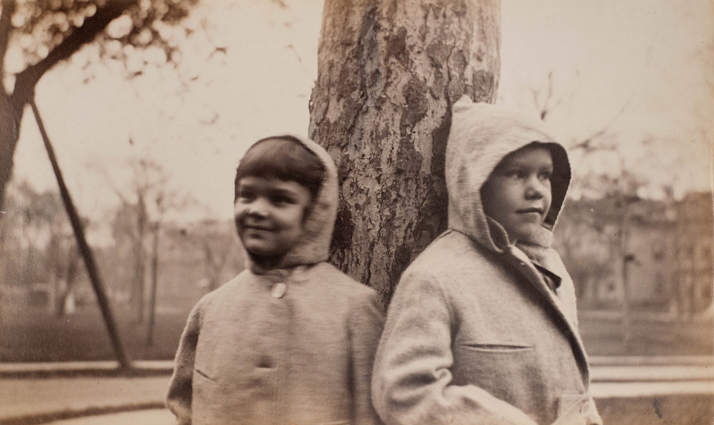 Sepia tone photograph of two children in hooded coats with the hoods up standing with their backs to a tree in a field. Trees and a house are out of focus in the background and each child looks off into the distance on their respective side, left and right. 